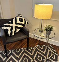 Dark gray Ikea NILS chair with black and white geometric pillow beside an adjustable height glass and chrome Eileen Gray style side table topped with a chrome table lamp, faux plant, and abalone dish, all on a black and white geometric kilim rug.