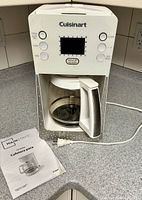 Front view of white Cuisinart drip coffee maker with digital display and control buttons. Manual placed beside it on counter.
