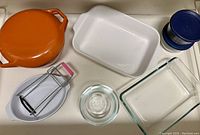 Overview of orange Copco lidded casserole, white baking dish, glass bowls, blue lidded Pyrex storage bowls, and a metal hand mixer with pink handle.