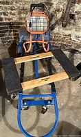 Photo showing blue handcart in flat position with padded black arms and wooden slats, orange worklight mounted on trolley behind.