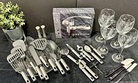 Overview of all kitchen utensils with metal handles and black grips, four wine glasses, corkscrew box, and other bar accessories on table.