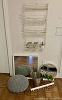 Collection arranged on floor against wall showing knitted pouf, square mirror, round mirror with shelf, wire caddy, steel waste basket, glass dispenser, and over door hanger.