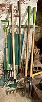 Photo showing six wooden-handled garden tools against a brick wall, including a pitchfork with green grip handle, post hole digger, hoe, and Fiskars tree branch pruner with green grips.