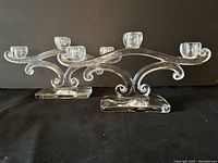 Pair of clear triad candleholders shown together front angle, highlighting shape and size, elegant curves and three candle cups.