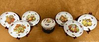 Top-down view of six Seltmann Weiden 7.5 inch hand-painted luncheon plates arranged in two rows surrounding a smaller Royal Worcester covered casserole bowl with fruit decorations on a brown surface.