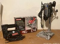 View of all three tools together: corded sabre saw on left, boxed LaserTrac laser level in center, drill press stand with rotary tool on right.