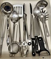 Drawer with stainless steel ladle, slotted spoon, tongs with black silicone tips, large serving spoon, balloon whisk, vegetable peeler, mesh strainer, soup spoons