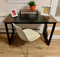 Desk with wood veneer edges and black inset work area, white molded plastic chair with chrome wire legs, small wood platform on desk with faux plant, two framed prints on desk edges.