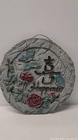 Front view of ceramic garden art plaque showing textured gray circular base with blue butterflies, red roses, green leaves, Asian character, and 'Happiness' written in cursive.
