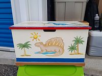 Front side of the wooden dinosaur toybox showing a tan spiked dinosaur, green palm trees, and a yellow sun on a cream background with red top trim.