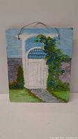 Front view of hand painted stone panel showing a white arched garden door with blue flowers, greenery and a stone path.