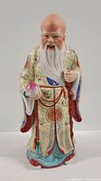 Front view of ceramic sculpture showing elderly man with white beard, holding a peach and staff, dressed in ornate robe.