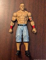 Front view of the John Cena WWE action figure showing muscular detail, light blue shorts with pockets, red and black armbands, and black shoes.