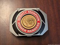 Front view of the Mighty Morphin Power Rangers morpher toy showing the central gold tiger emblem within a red circle and silver-gray casing with black details.