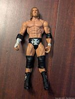 Front view of Mattel Triple H WWE action figure approximately 7 inches tall, showing muscular physique, black trunks with silver design, knee and wrist pads, and boots.
