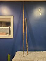 Two long handled antique tools with wooden handles and metal ends leaning against a blue wall.