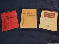 Three vintage typing books lying flat on cloth surface: a red "Typewriting and Office Practice" by Pitman, a tan "New Course in Typewriting" by Pitman, and a beige "High School Typewriting" by Roszell and Hewitt published by Gregg.