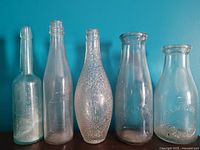 Photo showing 5 antique glass bottles with various shapes and embossed texts including ribbed and textured surfaces.
