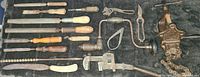 Photo showing a collection of various antique hand tools laid out on a fabric surface, including files, a saw, pliers, hand braces, and a chain tool.