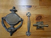 Photo shows three antique/vintage door hardware items: an oval door knocker with ornate arms, a curved handle, and a lock mechanism with latch.