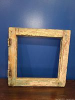 Square wooden window frame with faded paint and hinges leaning against a blue wall