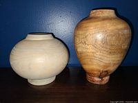Two handmade wooden vases, one light and round, one darker and taller, displayed against blue wall