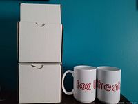 Two white coffee mugs with 'fox theater' printed in red text are shown next to two stacked white boxes likely used for packaging.