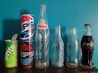 Six vintage soda containers against blue wall. Three Pepsi cans with colorful designs, one Sprite can with rust, two clear glass Pepsi bottles, one clear glass Coca-Cola bottle.