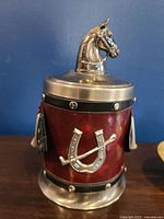 Front side showing horse head lid, red textured body with metal horseshoe and riding crop decoration, metal base and trim