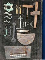 Photo of assorted vintage tools and hardware laid out on black fabric, showing corkscrew, scraper, wooden mallet/plane tool, metal hooks, weights, wrenches, and other metal fittings.