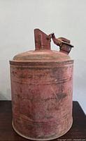 Side view of the vintage red metal gas can showing spout and handle with rust and wear.