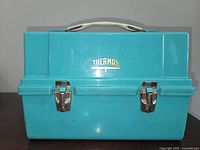 Closed front view of teal blue vintage Thermos cooler with two metal latch clasps and a white handle.