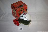 Photo showing white Steve Canyon helmet with green sun visor and red mouthpiece alongside its original box with orange graphics and text.