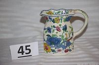 Full view of the ceramic hand-painted pitcher with colorful floral pattern and handle showing the face motif.