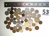 Top-down photo of 53 mixed foreign coins arranged under a ruler showing sizes up to about 25 mm, laid out flat with various designs and metals.