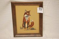 Frontal view of framed painting of a fox sitting on ground with rocks and grass, framed in wood, with lot number tag.