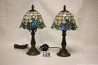 Pair of small stained glass table lamps with ornate metal bases, both lamps plugged into power cords, beige background