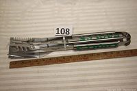 Three stainless steel BBQ utensils—from left to right: spatula with serrated edges and bottle opener, meat fork, and tongs—laid parallel with measuring ruler showing approx. 18 inches length.