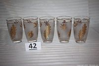 Front view of five frosted Libbey highball glasses with gold foliage decoration and gold rims, arranged in a row on striped fabric background.