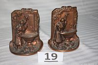 Pair of vintage brass or bronze bookends depicting a woman drinking at a fountain with a lion head spout, placed side by side on a light-colored fabric surface with lot number '19' shown in front.