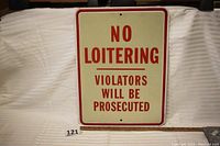 Front view of a rectangular vintage steel sign with red text 'NO LOITERING VIOLATORS WILL BE PROSECUTED' on white background, placed against a white cloth with a wooden ruler below for scale.