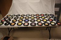 Table covered with a white cloth and arranged with more than 40 vintage 45 RPM records in rows, showing colorful labels and black vinyl discs from the 1960s-70s era.