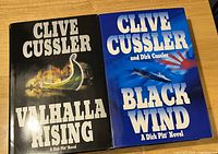 Two hardcover Clive Cussler novels: 'Valhalla Rising' with a Viking ship graphic, and 'Black Wind' featuring a jet aircraft and rising sun on blue background.