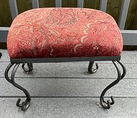 Square footstool with red patterned fabric upholstery and curved black metal legs shown on a metal surface outdoors.