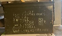 Side view of green metal ammunition box showing the stenciled white text indicating 100 cartridges of 50 caliber ball ammunition with lot number and other markings.