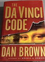 Front cover of The Da Vinci Code hardcover book by Dan Brown showing the red dust jacket with gold text and Mona Lisa partial image.