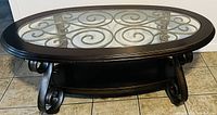 Front angle view showing the dark wood frame oval shaped coffee table with transparent tempered glass inset and dark metal scrollwork beneath, supported by curved powder-coated metal legs.