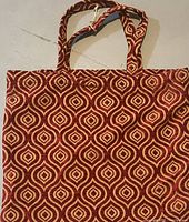 Front view of a red and beige patterned velour carry bag with two shoulder straps laid flat on surface.