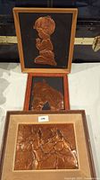 Three copper embossed plaques arranged on a white surface, displaying a child praying, a horse's head, and two horses with riders.