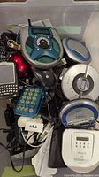 Bin filled with assorted electronics including multiple portable CD players, handheld calculators, digital cameras, and cables.